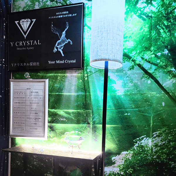 Yクリスタル探偵社のオフィス内観。白銀の額に入った探偵の標識、社名と「Your Mind Crystal」が記された浮き出し式内看板、そしてダイヤモンドのオブジェが間接照明に照らされ、幻想的な雰囲気を醸し出している。