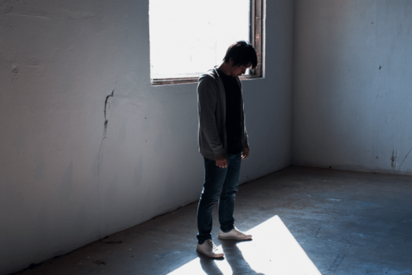 窓からの光が差し込む薄暗い部屋で、うつむいて立ち尽くす一人の日本人男性の姿。ストーカー行為や不安な日々を送る人物の孤独感や不穏な状況を表現しています。