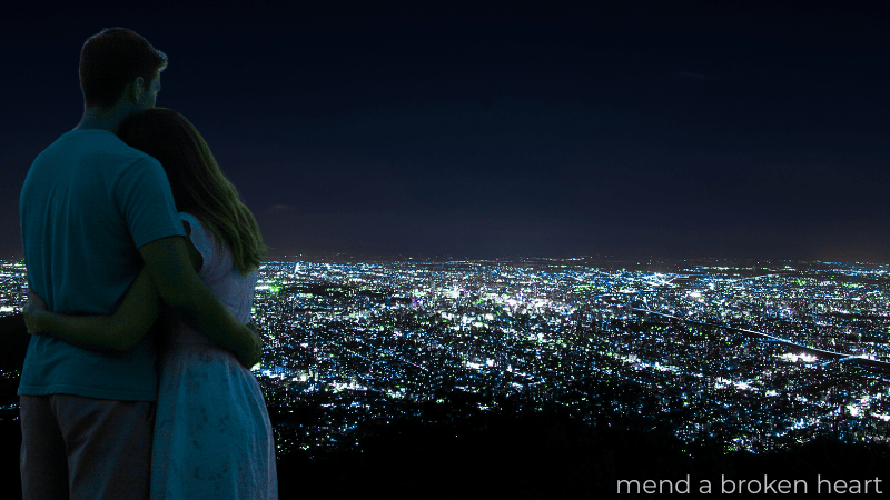 夜景が広がる丘の上で、肩を寄せ合い遠くを見つめるカップルの後ろ姿。関係修復によって得られる「新しい未来」と「希望」を象徴するイメージ。「mend a broken heart」の文字が小さく添えられている。