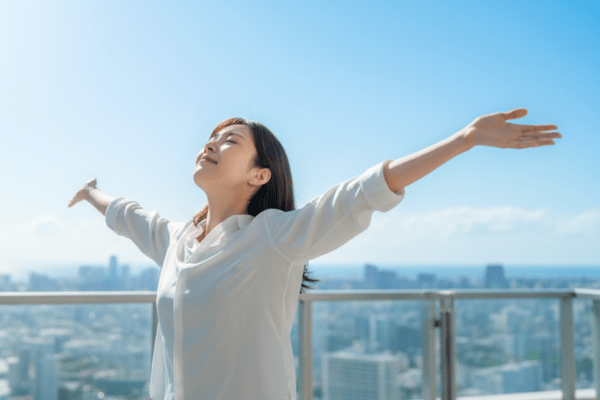 青空の下、バルコニーで両手を広げ、開放感と喜びを感じる女性の姿。困難を乗り越えた新しい未来の象徴。