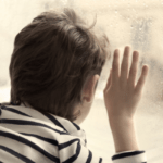 雨の降る窓の外を一人で見つめる子供の背中。孤独や不安を抱える子供の心の痛みと、それを救い出し笑顔を取り戻すというYクリスタル探偵社の使命を象徴するイメージ画像。